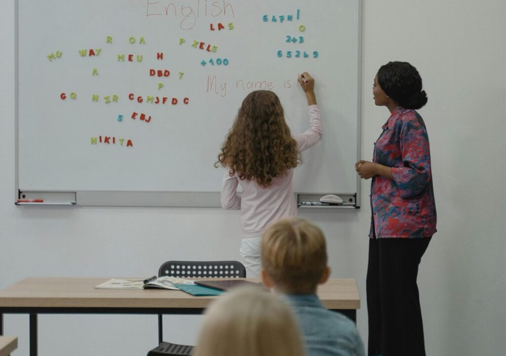 Teacher instructing diverse students with magnetic letters on a classroom whiteboard.